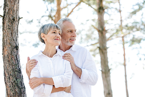 smiling senior couple in white shirts embracing and looking away in forest