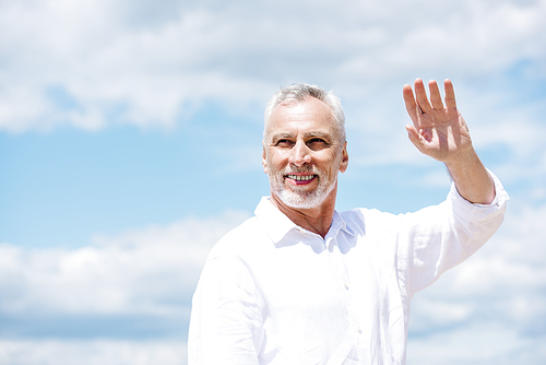 smiling senior man with beard looking away and waving hand under blue sky in sunny day