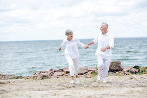 full length view of happy senior couple holding hands and looking at each other while running at beach