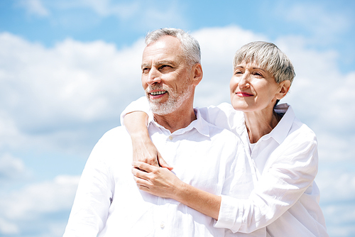 happy senior couple in white shirts embracing under blue sky