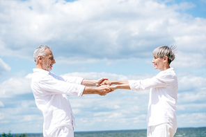side view of happy smiling senior couple holding hands and looking at each other near river