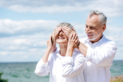 smiling senior man covering eyes for wife near river in sunny day