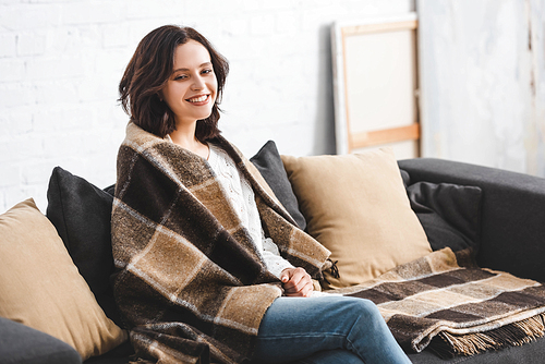 happy brunette woman in blanket sitting on sofa in cozy living room