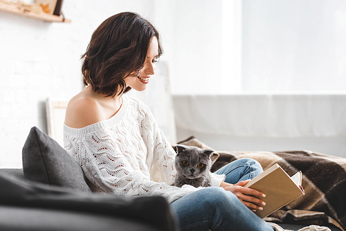 attractive smiling woman reading book on sofa with scottish fold cat
