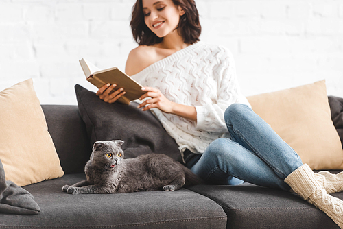 brunette smiling woman reading book with scottish fold cat on sofa