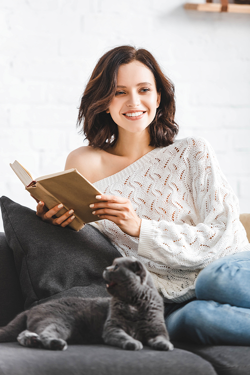 smiling woman reading book with scottish fold cat on sofa