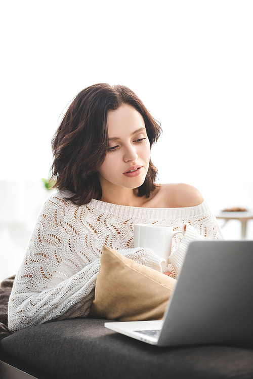 beautiful pensive girl with cup of coffee using laptop at cozy home