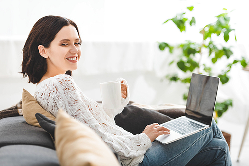 cheerful girl with cup of tea using laptop at cozy home