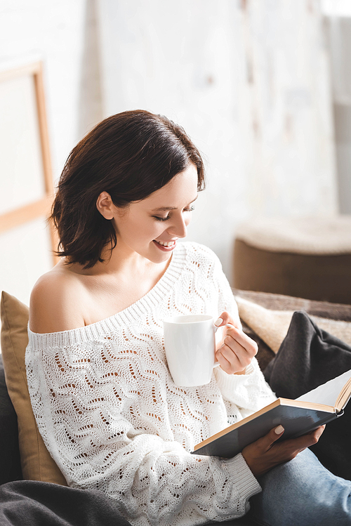 cheerful brunette girl reading book on sofa with coffee cup