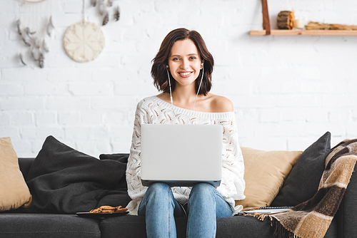 smiling girl using earphones and laptop on sofa with cookies