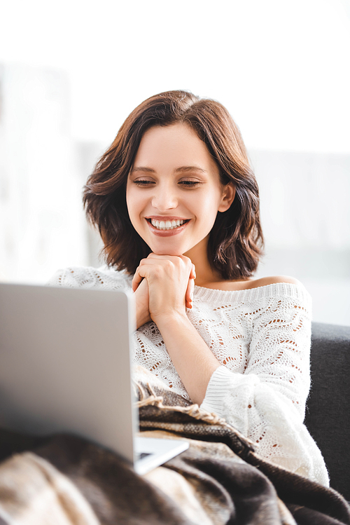 attractive smiling girl in blanket using laptop on sofa in cozy living room