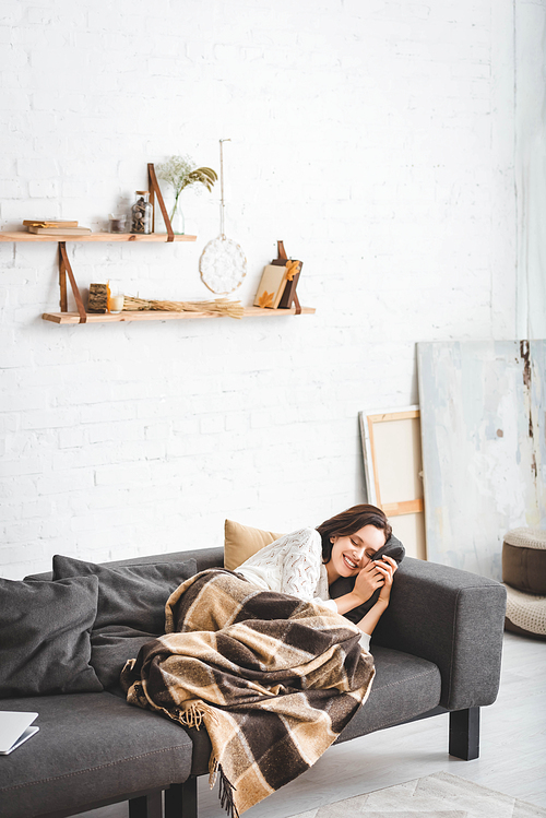happy woman with closed eyes relaxing in blanket in cozy living room