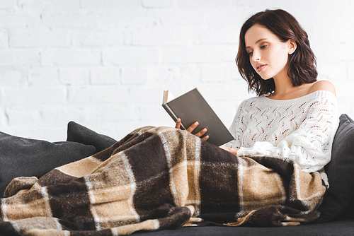 beautiful young woman in blanket reading book on sofa at home
