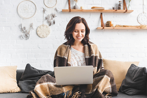 beautiful cheerful girl in blanket using laptop in living room with dream catchers
