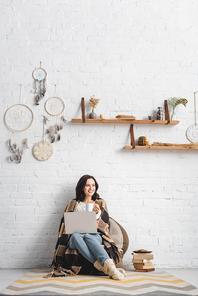 cheerful brunette girl with cookies and coffee using laptop in living room with dream catchers