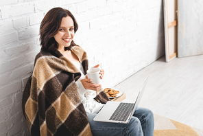 beautiful smiling girl in blanket with cookies and coffee using laptop