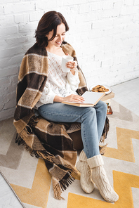 happy girl in blanket reading book in cozy living room with coffee and cookies