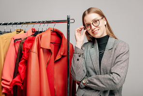 happy stylist touching glasses near trendy clothing isolated on grey