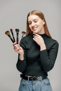 happy makeup artist holding cosmetic brushes isolated on grey