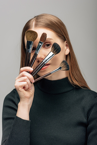 young makeup artist covering face with cosmetic brushes isolated on grey