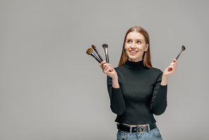 cheerful makeup artist holding cosmetic brushes isolated on grey