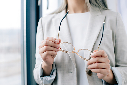 cropped view of executive businesswoman in grey suit holding eyeglasses in office