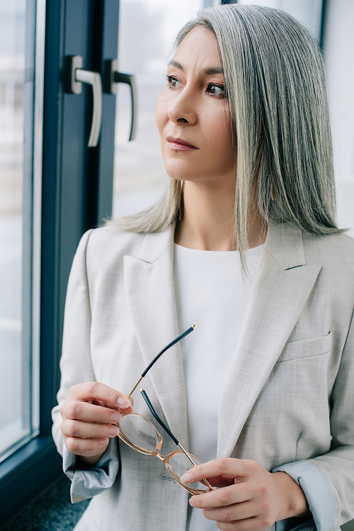 beautiful asian businesswoman with grey hair in grey suit holding eyeglasses and looking through window