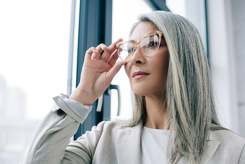 thoughtful asian businesswoman with grey hair in grey suit and eyeglasses in office