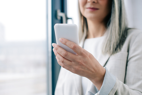 cropped view of businesswoman with grey hair using smartphone in office