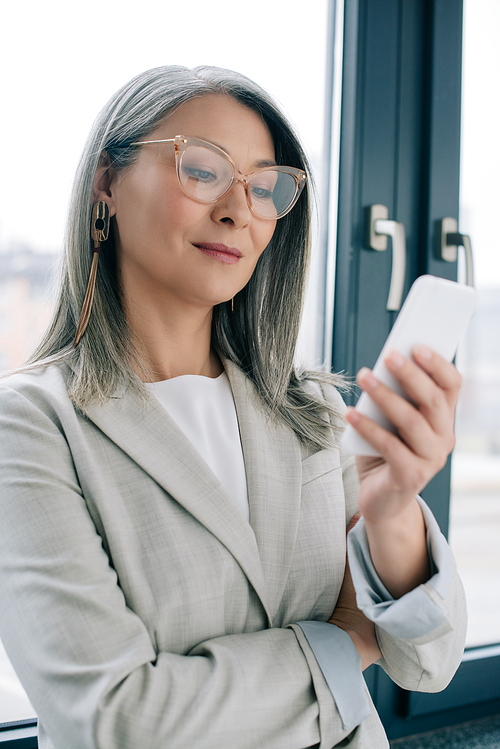 attractive asian businesswoman in eyeglasses with grey hair using smartphone in office