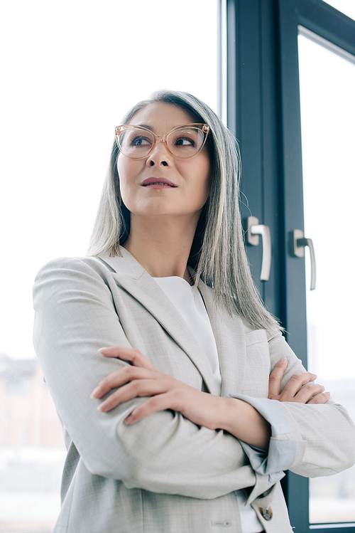 corporate asian businesswoman with crossed arms and grey hair standing in grey suit and eyeglasses in office