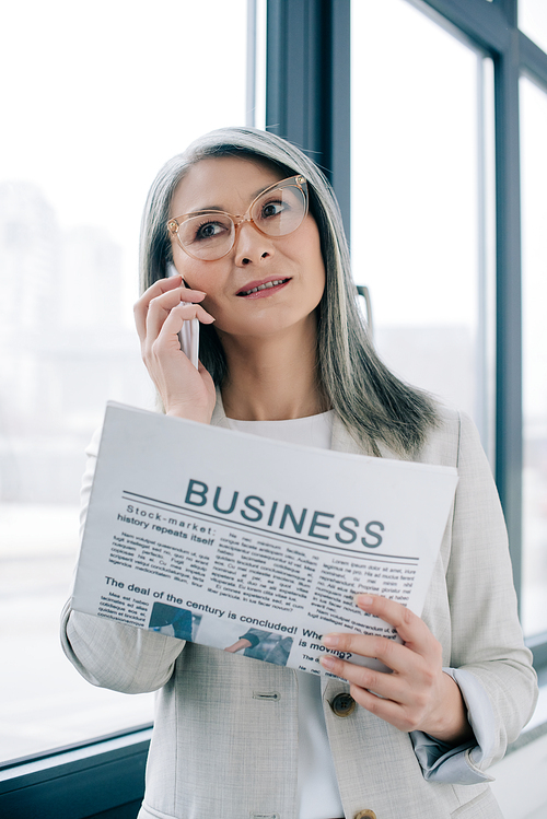 confident asian businesswoman in eyeglasses talking on smartphone and holding business newspaper in office