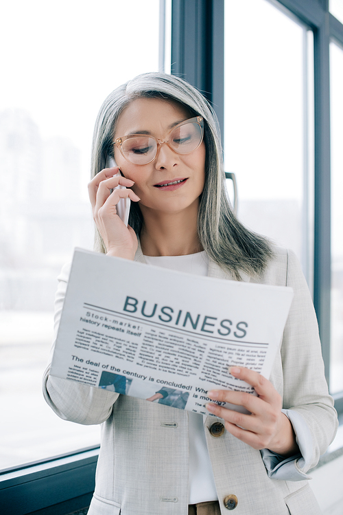 attractive asian businesswoman in eyeglasses talking on smartphone while reading business newspaper in office
