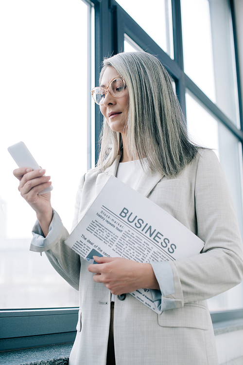 thoughtful asian businesswoman in eyeglasses using smartphone and holding business newspaper in office