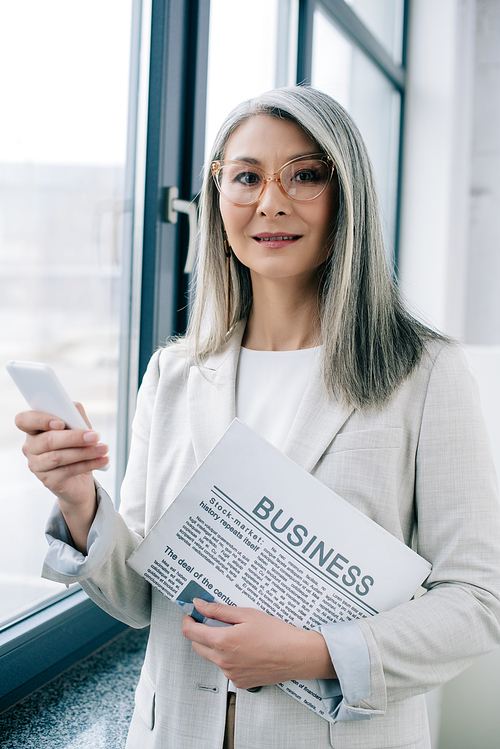 beautiful asian businesswoman in eyeglasses using smartphone and holding business newspaper in office