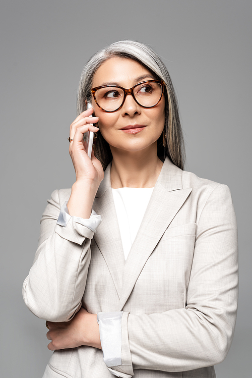 professional asian businesswoman in eyeglasses with grey hair talking on smartphone isolated on grey