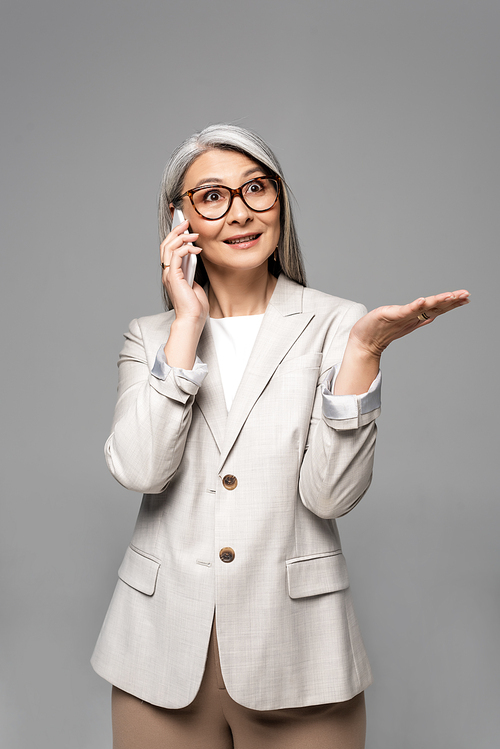 surprised asian businesswoman in eyeglasses with grey hair talking on smartphone isolated on grey