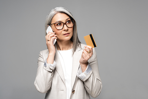 beautiful asian businesswoman with grey hair talking on smartphone and holding credit card isolated on grey