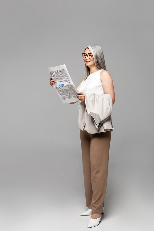 attractive asian businesswoman in eyeglasses reading business newspaper on grey