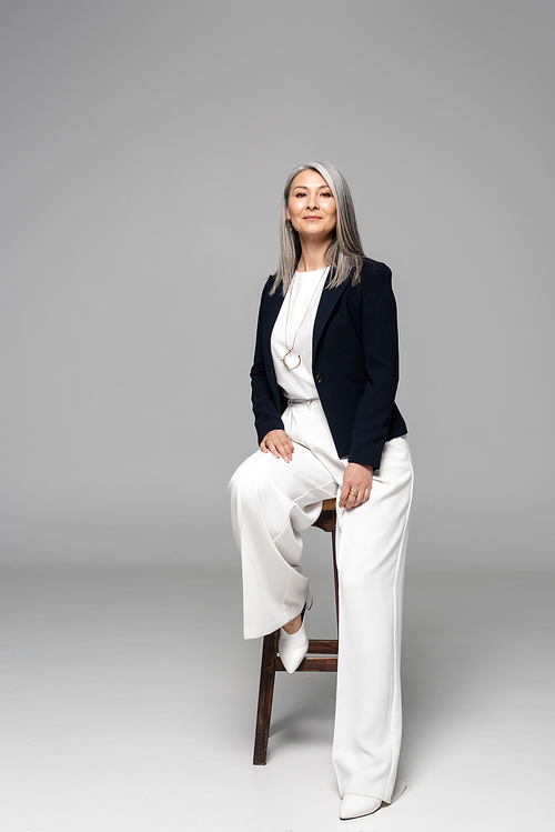 asian stylish businesswoman with grey hair sitting on stool on grey