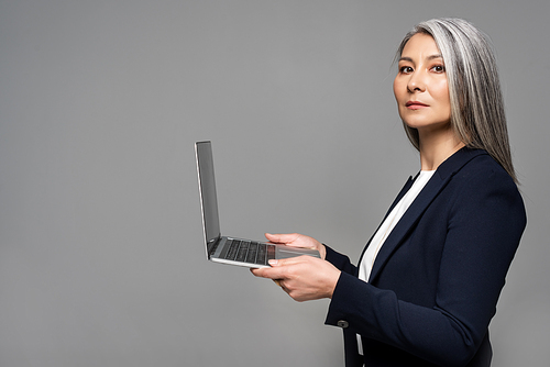 attractive serious asian businesswoman with grey hair using laptop isolated on grey