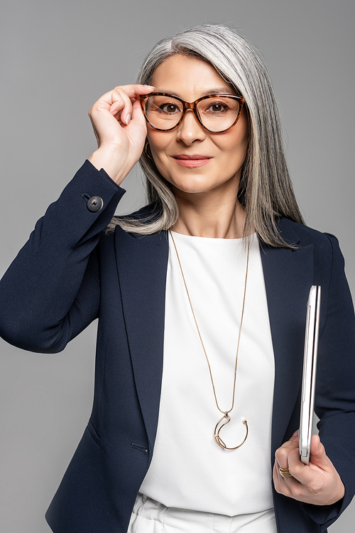 smiling asian businesswoman with grey hair holding laptop isolated on grey