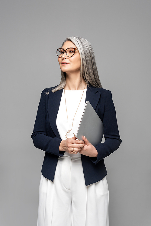 confident asian businesswoman with grey hair holding laptop isolated on grey