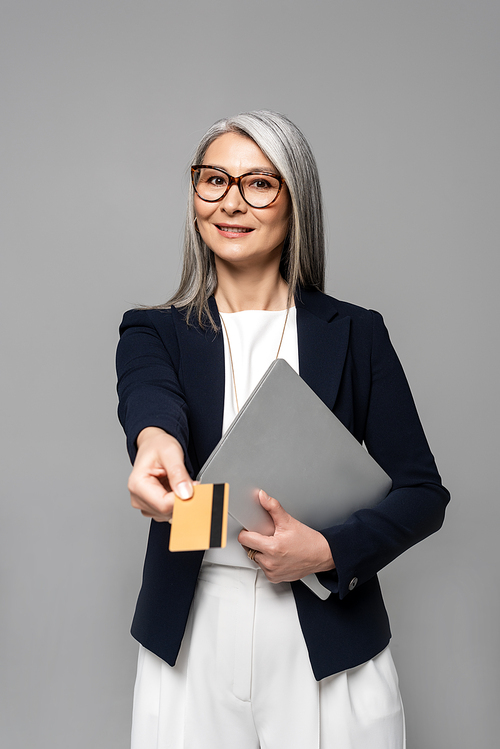 smiling asian businesswoman with grey hair shopping online with credit card and laptop isolated on grey