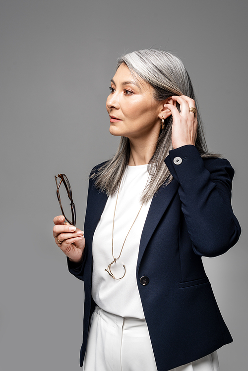 asian businesswoman with grey hair holding eyeglasses isolated on grey