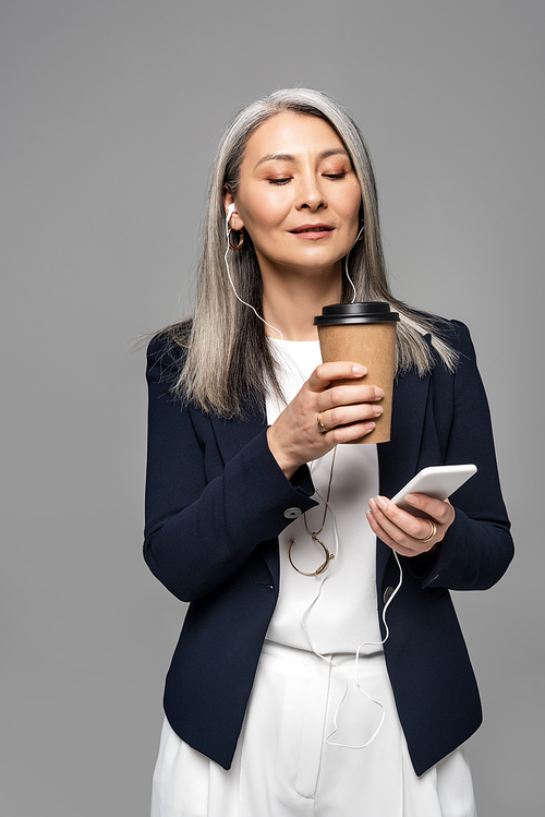 beautiful asian businesswoman with coffee to go listening music with earphones and smartphone isolated on grey