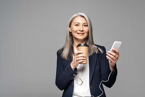 emotional asian businesswoman with coffee to go listening music with earphones and smartphone isolated on grey