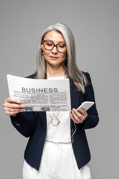 confident asian businesswoman in eyeglasses listening music with earphones and smartphone while reading business newspaper isolated on grey