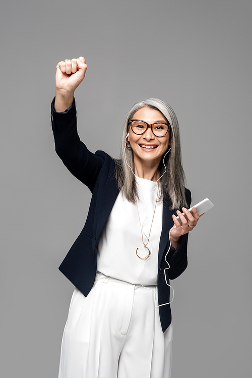 excited asian businesswoman with grey hair listening music with earphones and smartphone isolated on grey