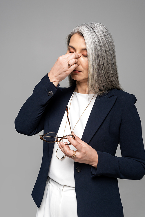 tired asian businesswoman with grey hair holding eyeglasses isolated on grey